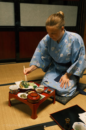 Woman in a blue floral kimono sitting on the floor, eating a meal with chopsticks.