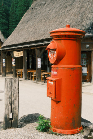 Red vintage-style postbox in front of a traditional building with a thatched roof.