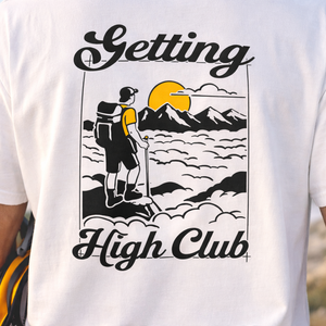 White t-shirt with a graphic design of a person hiking with mountains and text 'Getting High Club'.