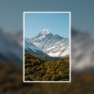 Snow-capped mountain peak with green forest below, framed by a white border. Features Mount Cook / Aoraki and nearby lakes in New Zealand.