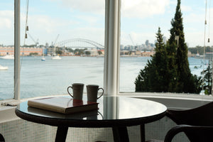 Round table with two cups and a book in front of large windows with a view of a waterfront.