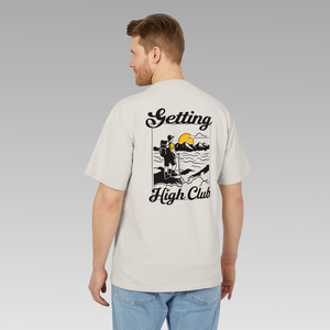 Man wearing a beige t-shirt with 'Getting High Club' hiking graphic and text on the back, standing against a gray background.