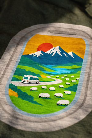 Graphic tee of a scenic landscape with mountains, sheep, and a van on a green fabric background showcasing New Zealand/Aotearoa.
